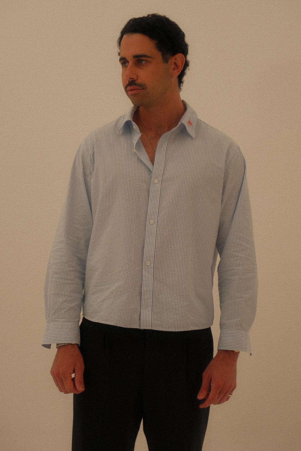 A man wearing a light blue and white vertically striped long-sleeve button-down shirt with black pants, standing against a plain beige background.