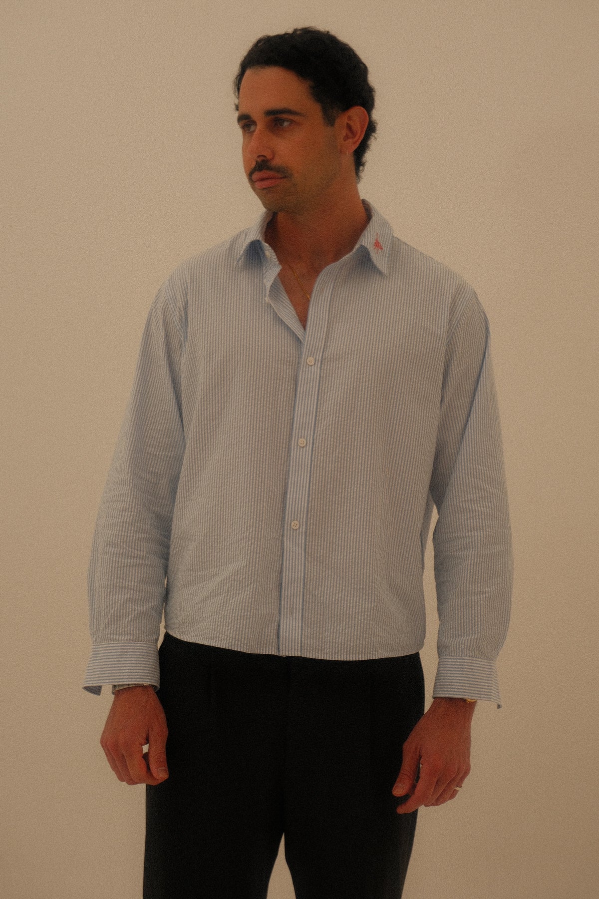 A man wearing a light blue and white vertically striped long-sleeve button-down shirt with black pants, standing against a plain beige background.